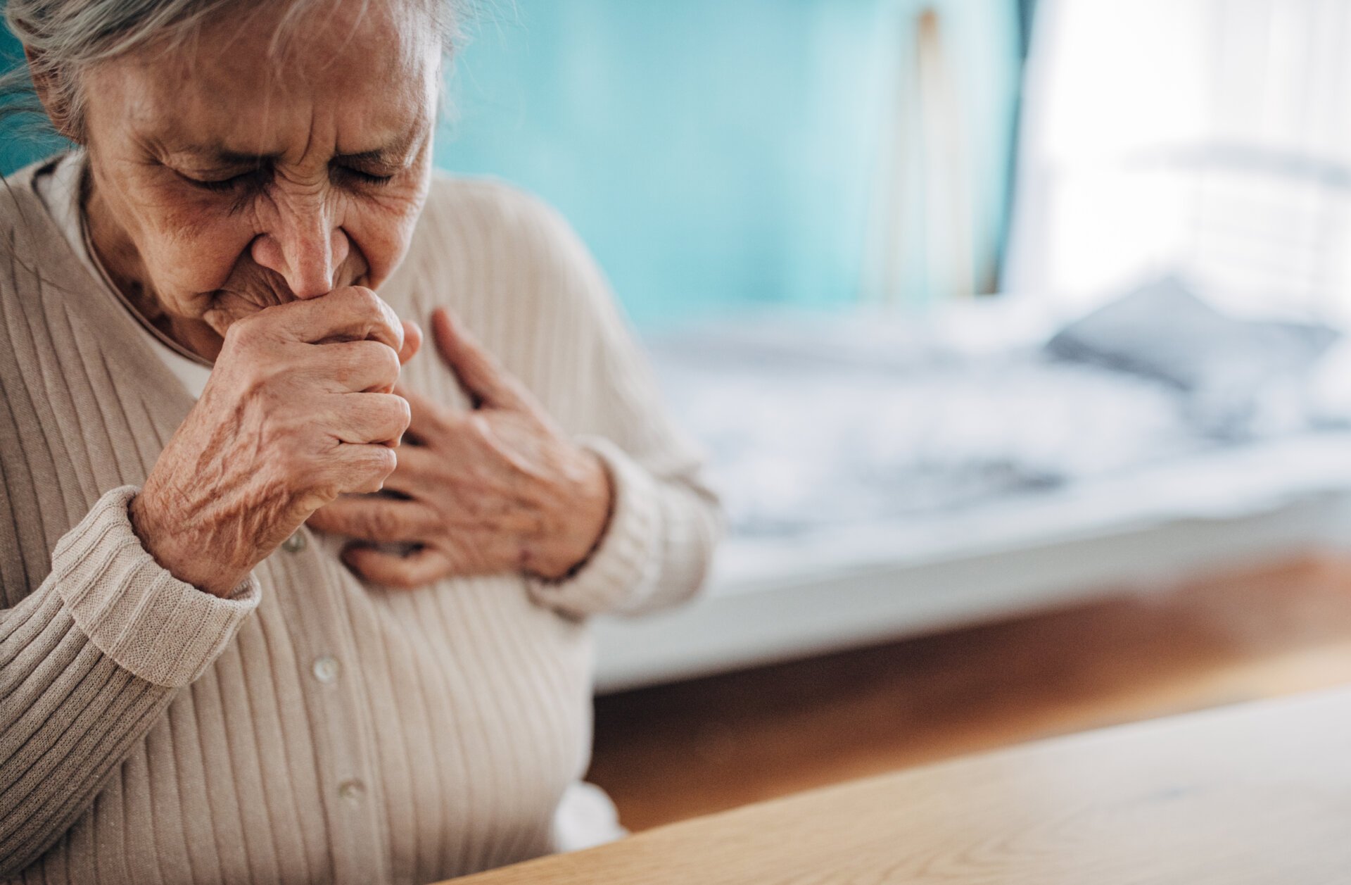 An elderly woman coughing and touching her chest from a respiratory illness.